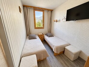 A compact room with two single beds, a small wooden dresser, a television mounted on the wall, and a window with curtains. - Cozy apt with direct slope access and shops, balcony, parking, pets ok! (Méribel)