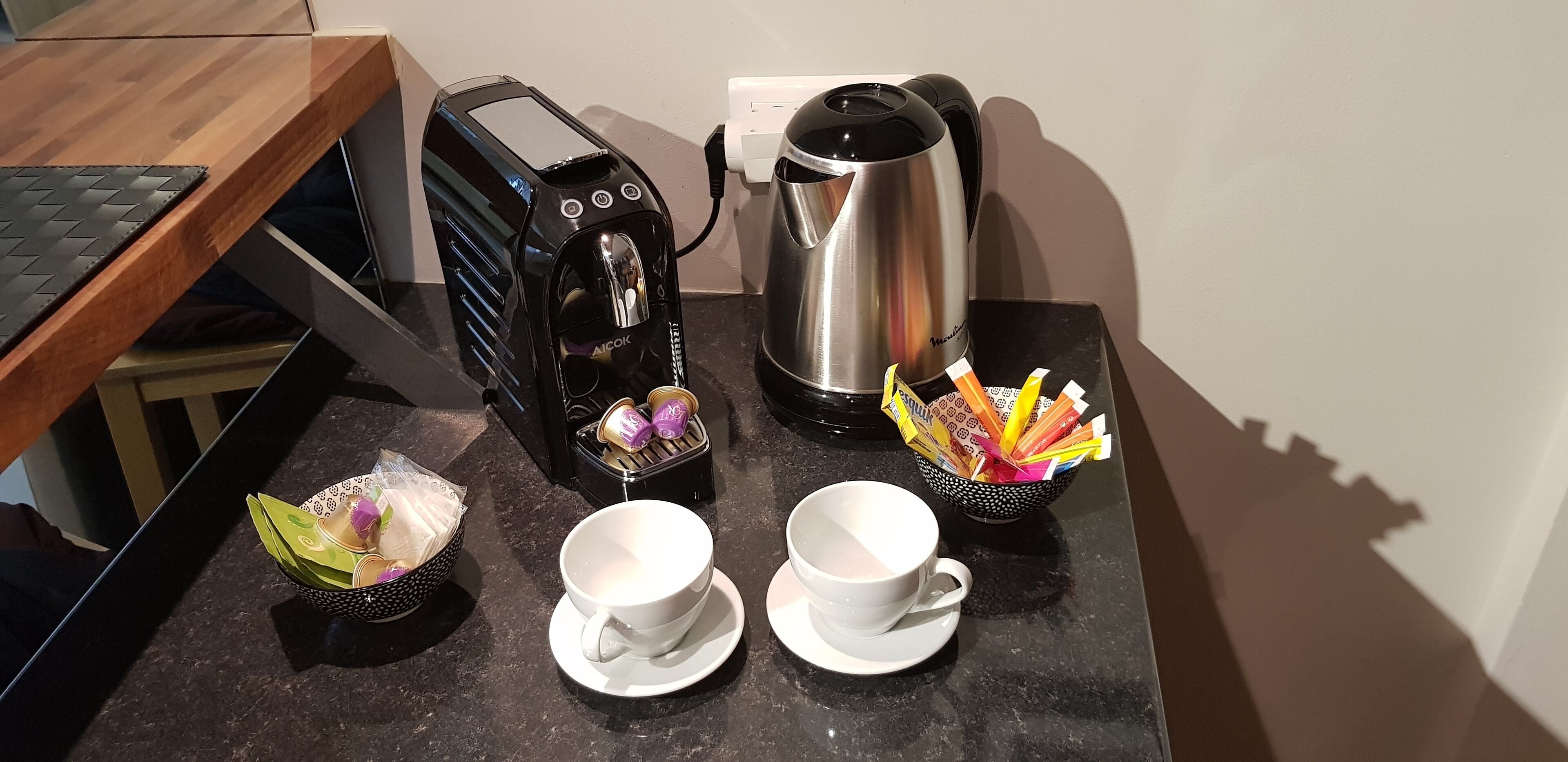 A kitchen counter with a toaster, a kettle, two cups, and a basket of tea bags.