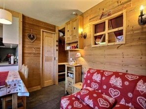 Quarto aconchegante com paredes de madeira, sofá vermelho com estampas de corações, uma mesa pequena e um armário com prateleiras.