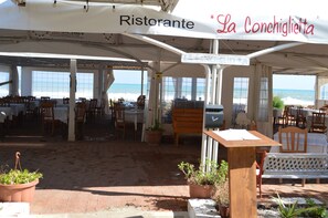 An outdoor dining area with tables and chairs, a wooden bench, and potted plants. - Il Veliero (Pachino)
