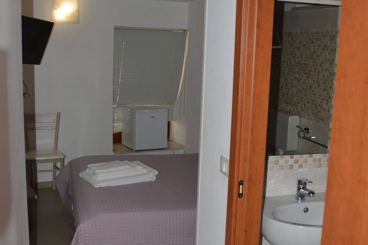 A narrow hallway leading to a bedroom with a bed, a chair, and a small refrigerator. A bathroom with a sink and mirror is visible through an open door.