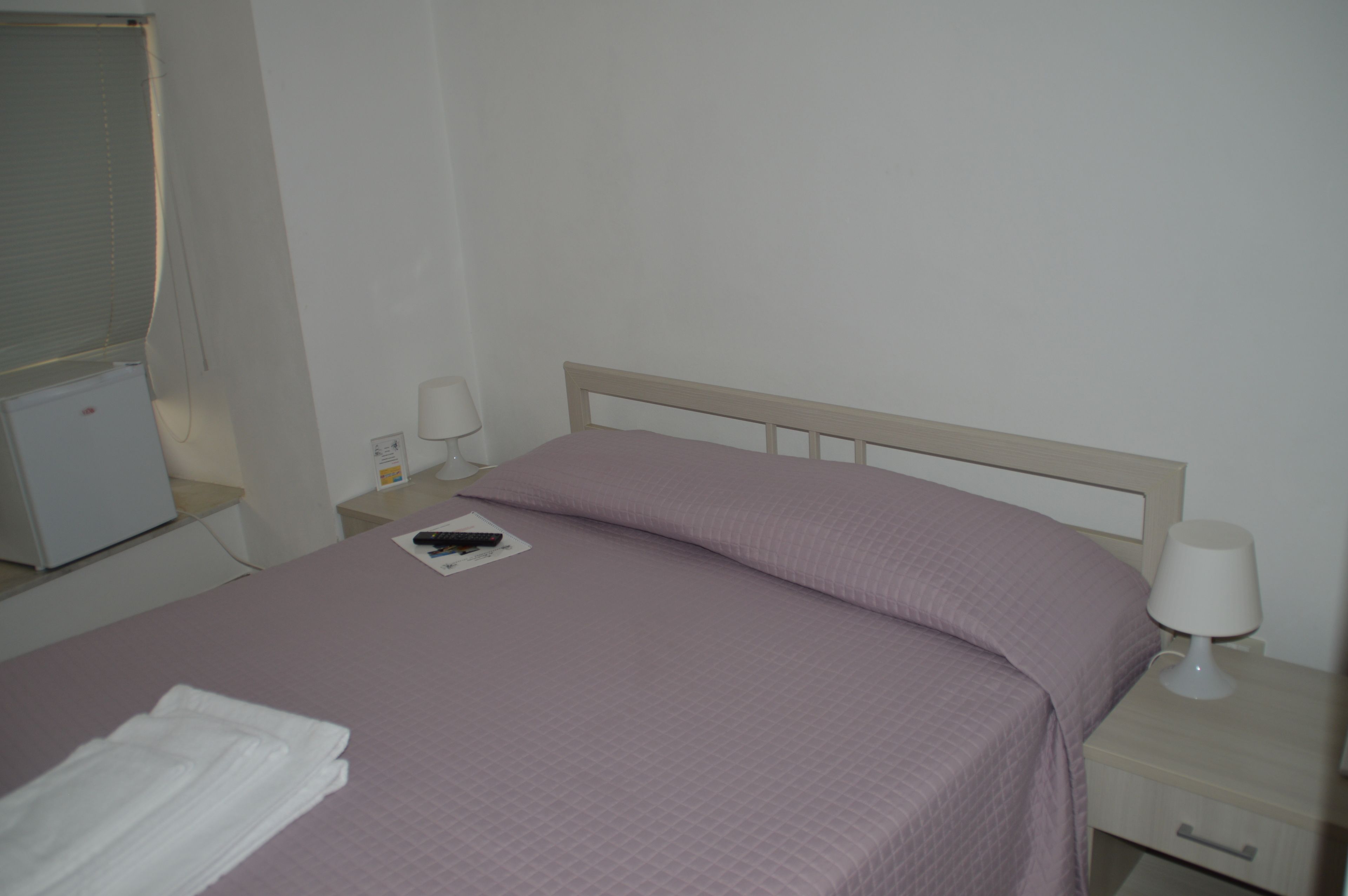 Una cama bien hecha con cabecera blanca, una lámpara de noche y una mesita con un control remoto y una caja de pañuelos.
