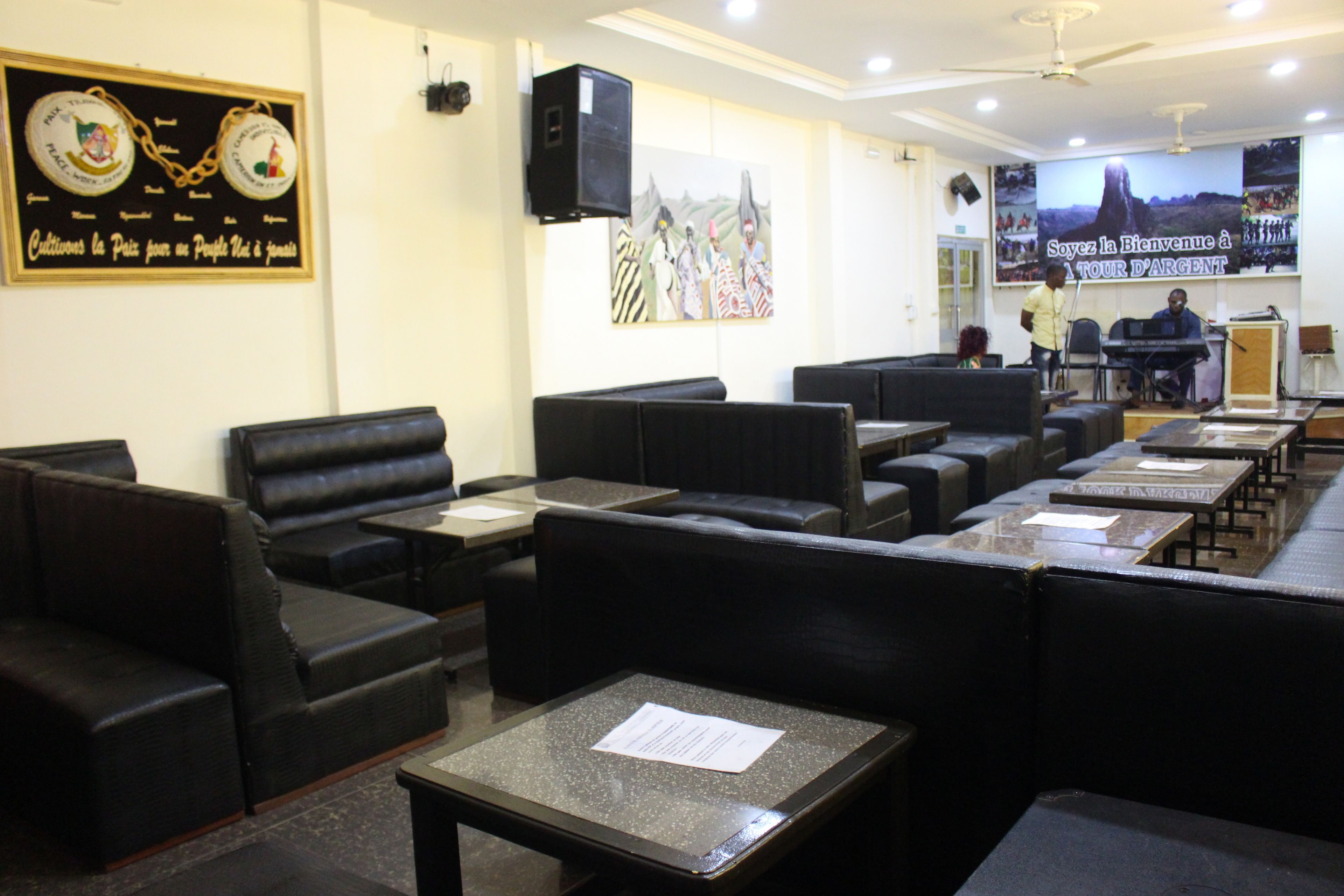 A restaurant with black leather booths and tables, a stage with a piano, and a large mural on the wall.