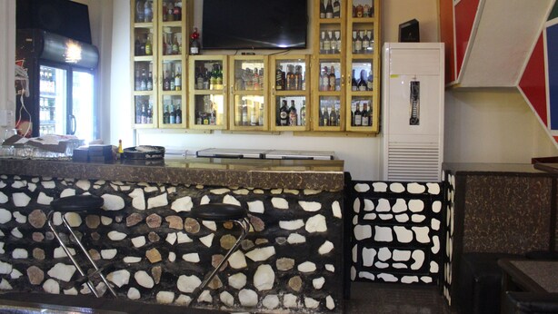 A bar area with a stone counter, stools, a television mounted above the bar, and shelves with bottles. - Hôtel la Tour d'Argent (Garoua)