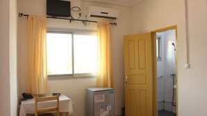 A small room with a table, a chair, a refrigerator, and a door leading to a bathroom. - Hôtel la Tour d'Argent (Garoua)