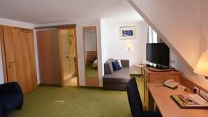 A hotel room with a sofa, a desk with a TV, a phone, and a small table with a lamp. There is a mirror and a door leading to a bathroom. - Zollersteighof (Albstadt)