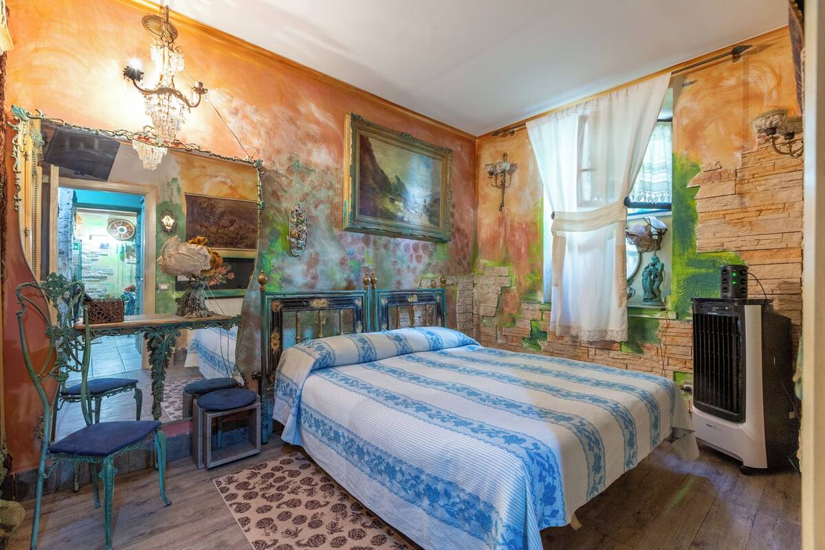Ein Schlafzimmer mit einem Bett, einem Schreibtisch und einem Stuhl. Es gibt ein Fenster mit Vorhängen und eine Wandheizung.