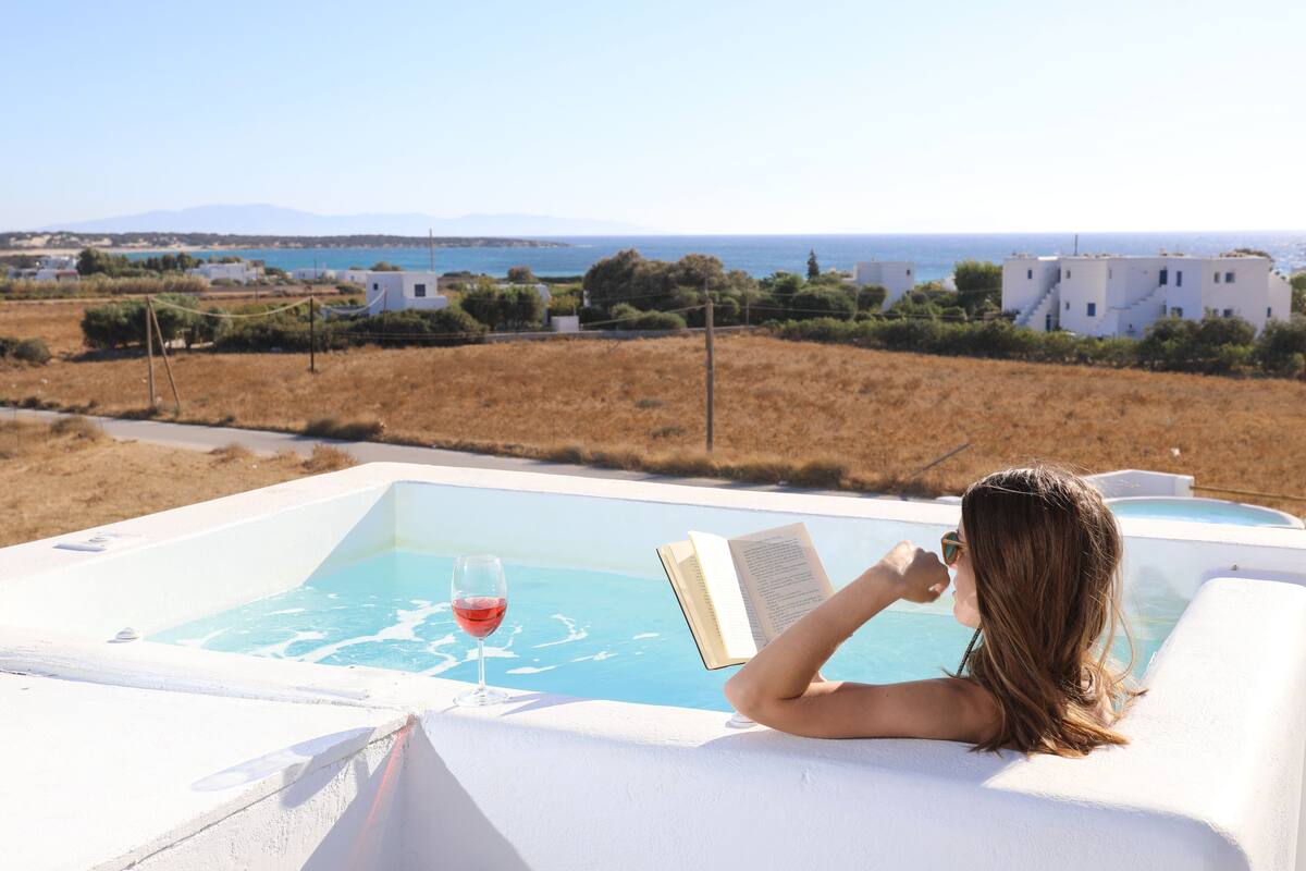 A person relaxing by a pool with a book and glass of wine, overlooking a coastal landscape with buildings and a clear sky.