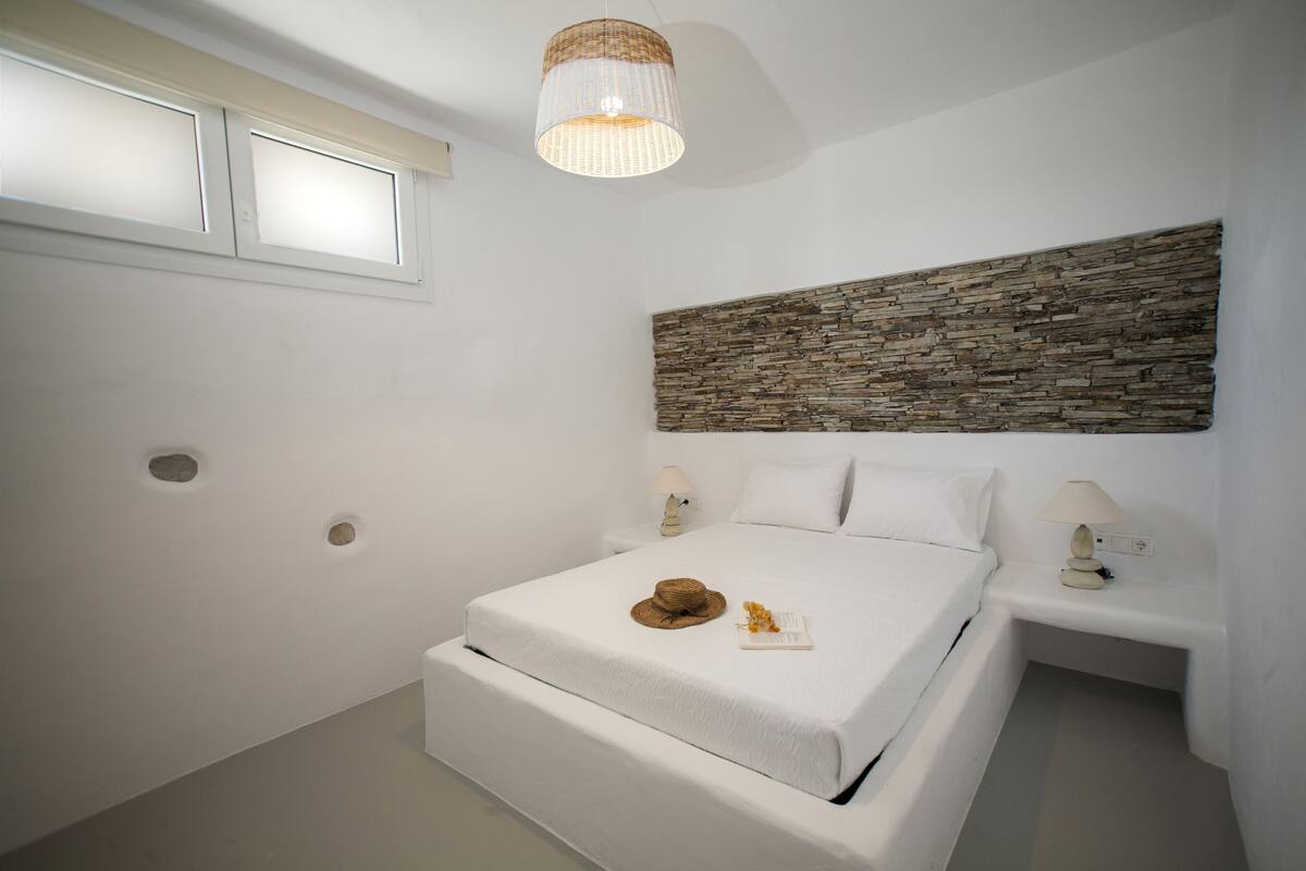 A bedroom with a large bed, two bedside tables, a stone accent wall, a woven light fixture, and a window with frosted glass.