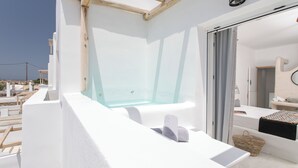 A modern, white-walled room with a glass-enclosed hot tub, a small bathroom with a sink and mirror, and a view of the sea. - Irida Vacation Suites (Naxos)