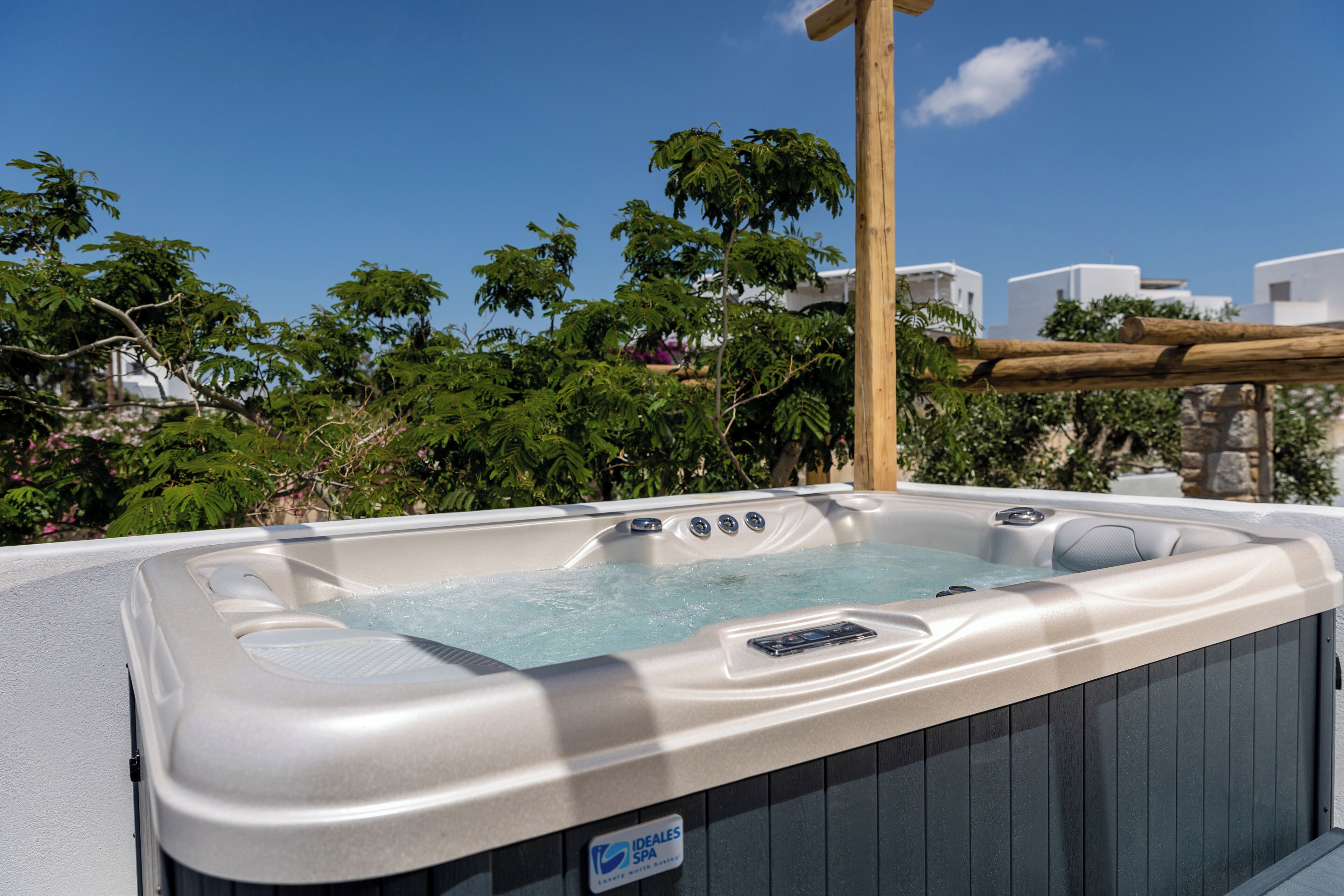 A hot tub with a wooden pergola overhead, surrounded by lush greenery and white buildings in the background.