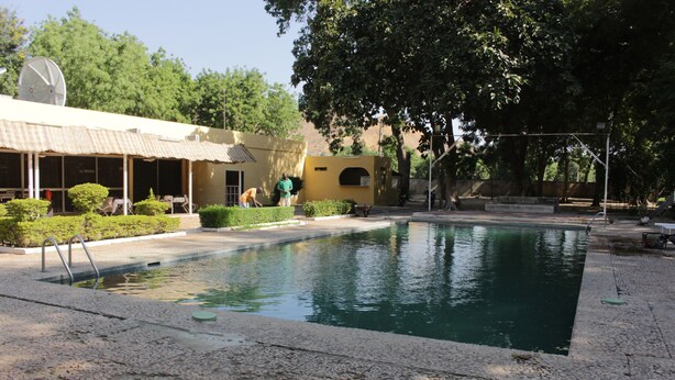 A swimming pool with a tiled deck, a satellite dish on a building, and a covered area with seating. - Hotel Mizao (Maroua)