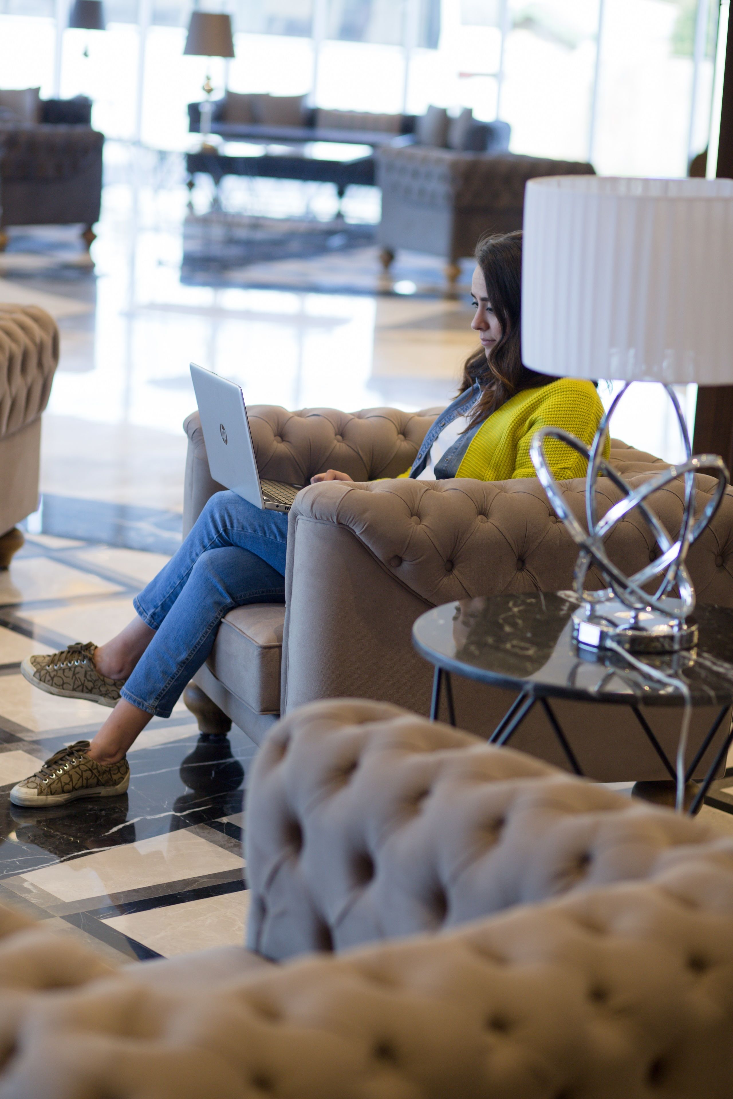 A person sitting on a tufted sofa with a laptop, in a well-lit lobby with modern furniture and a tiled floor.