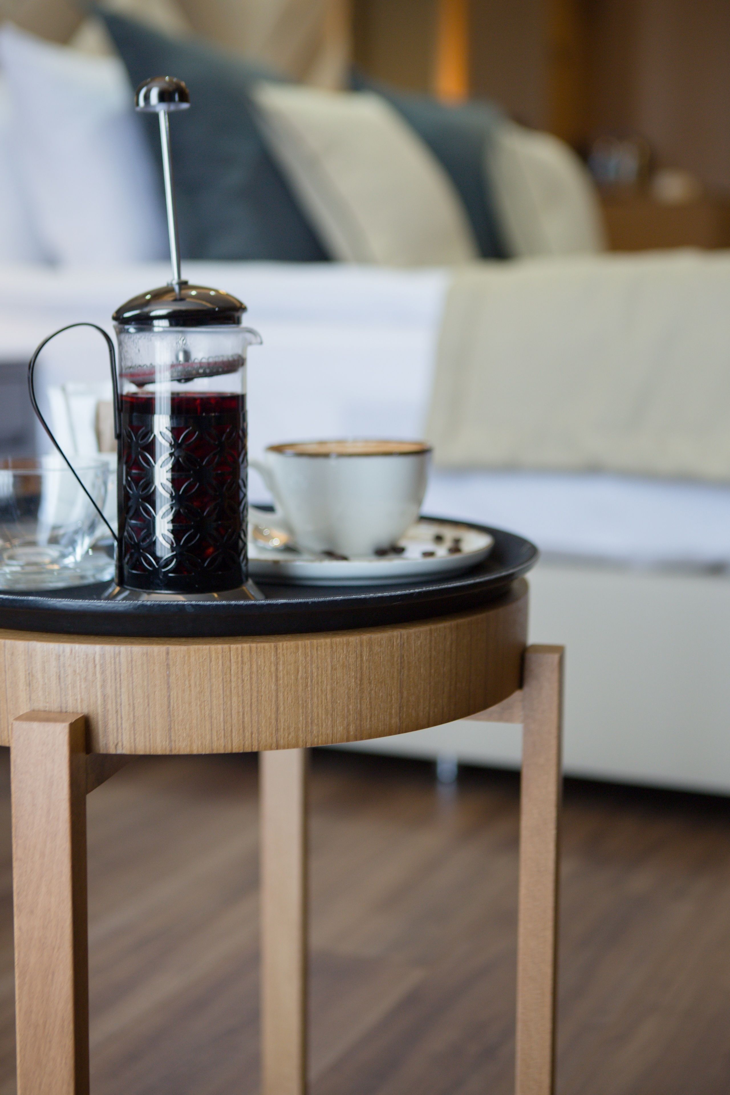 A coffee maker on a tray with a cup and saucer, a wooden table, and a bed with pillows in the background.