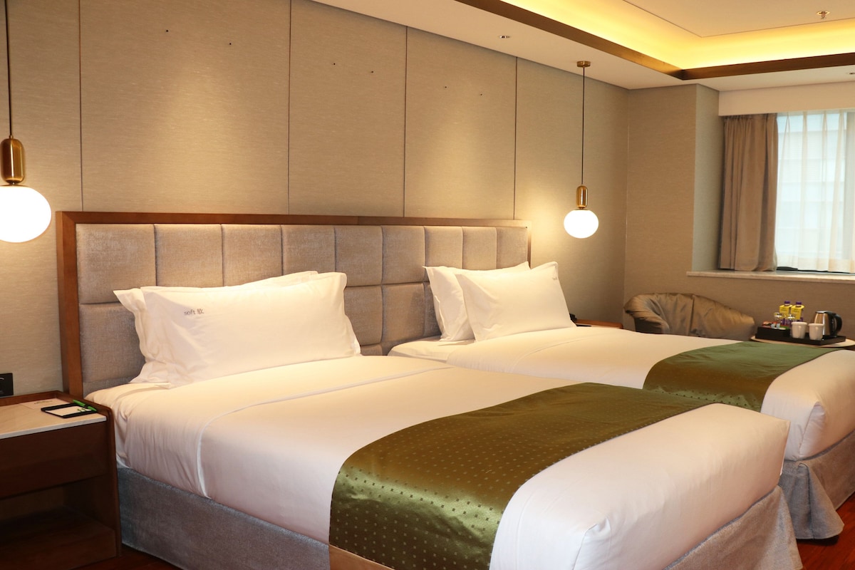 Een hotelkamer met twee bedden, een houten hoofdbord, een nachtkastje, een lamp, een raam met gordijnen en een klein tafeltje met een dienblad.
