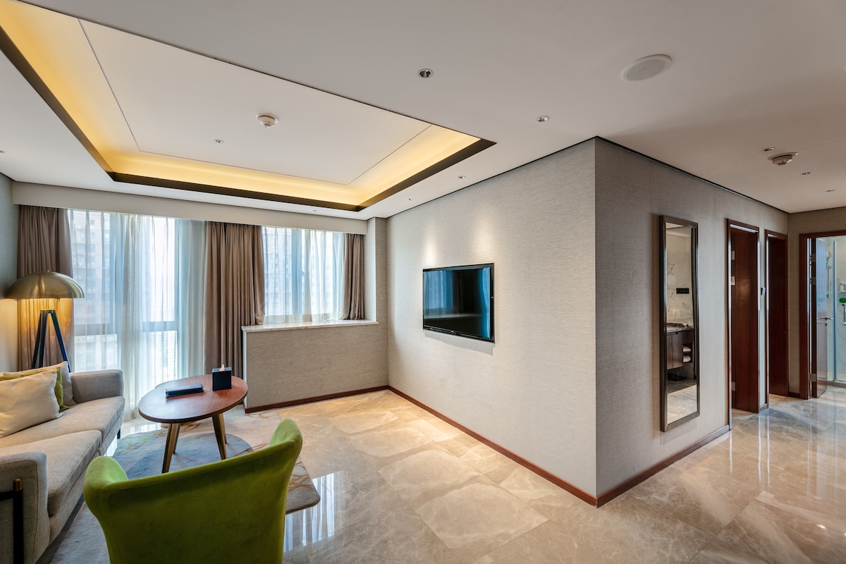 Een moderne hotelkamer met een bank, een groene fauteuil, een klein tafeltje, een flatscreen-tv aan de muur en een groot raam met gordijnen.