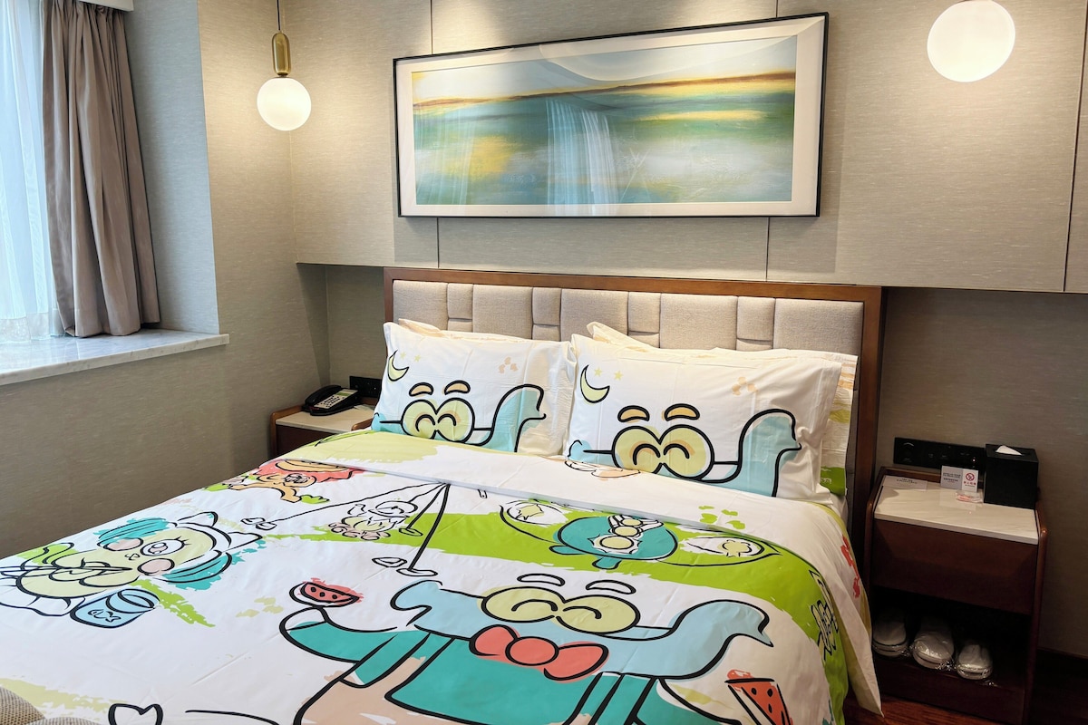 Een hotelkamer met een bed met beddengoed met tekenfilmfiguren, een nachtkastje met een telefoon, een schilderij boven het bed, en een schoenenrek.
