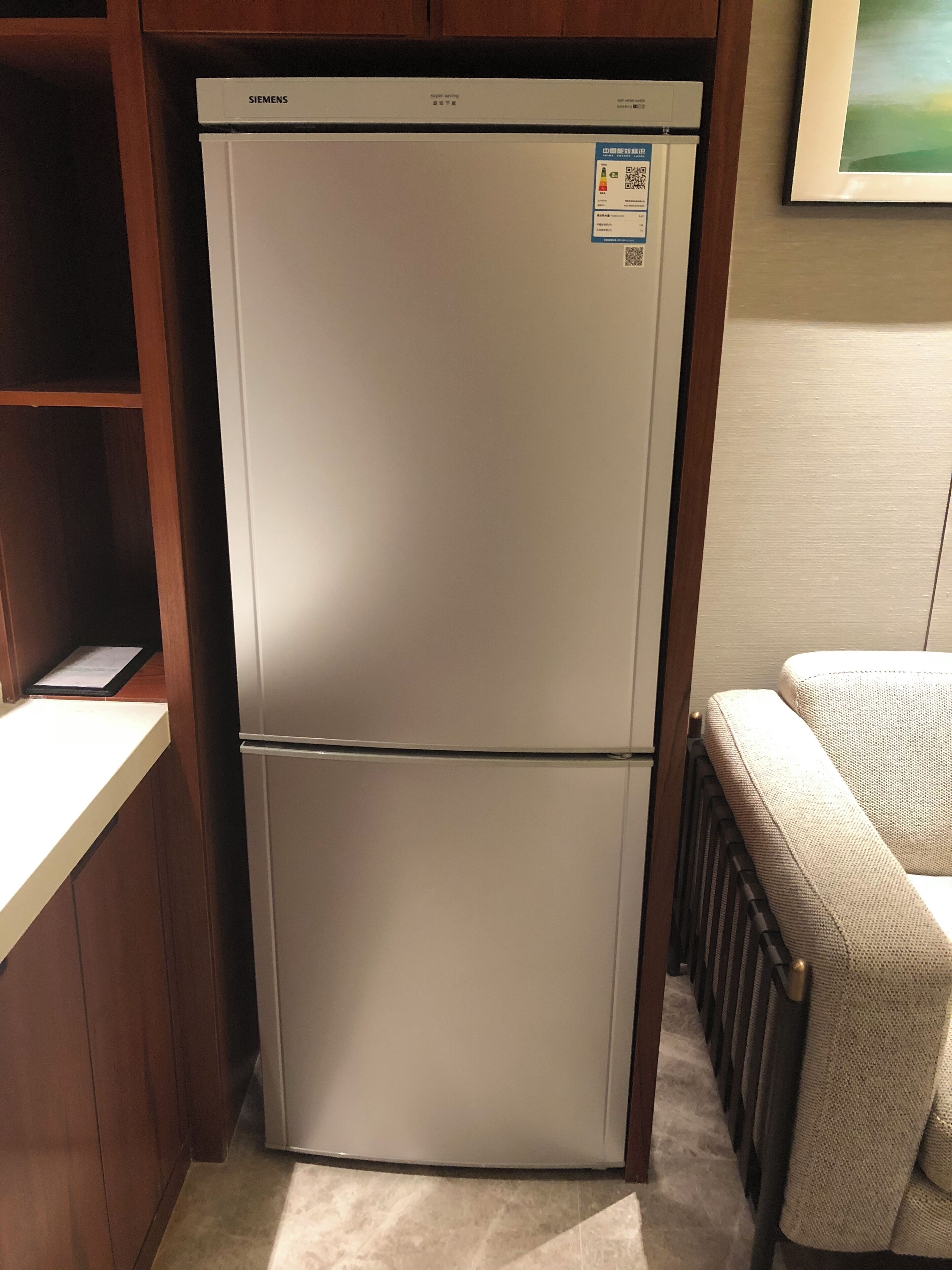 A white Siemens refrigerator with a digital display and a QR code sticker, located in a kitchen with wooden cabinets and a tiled floor.