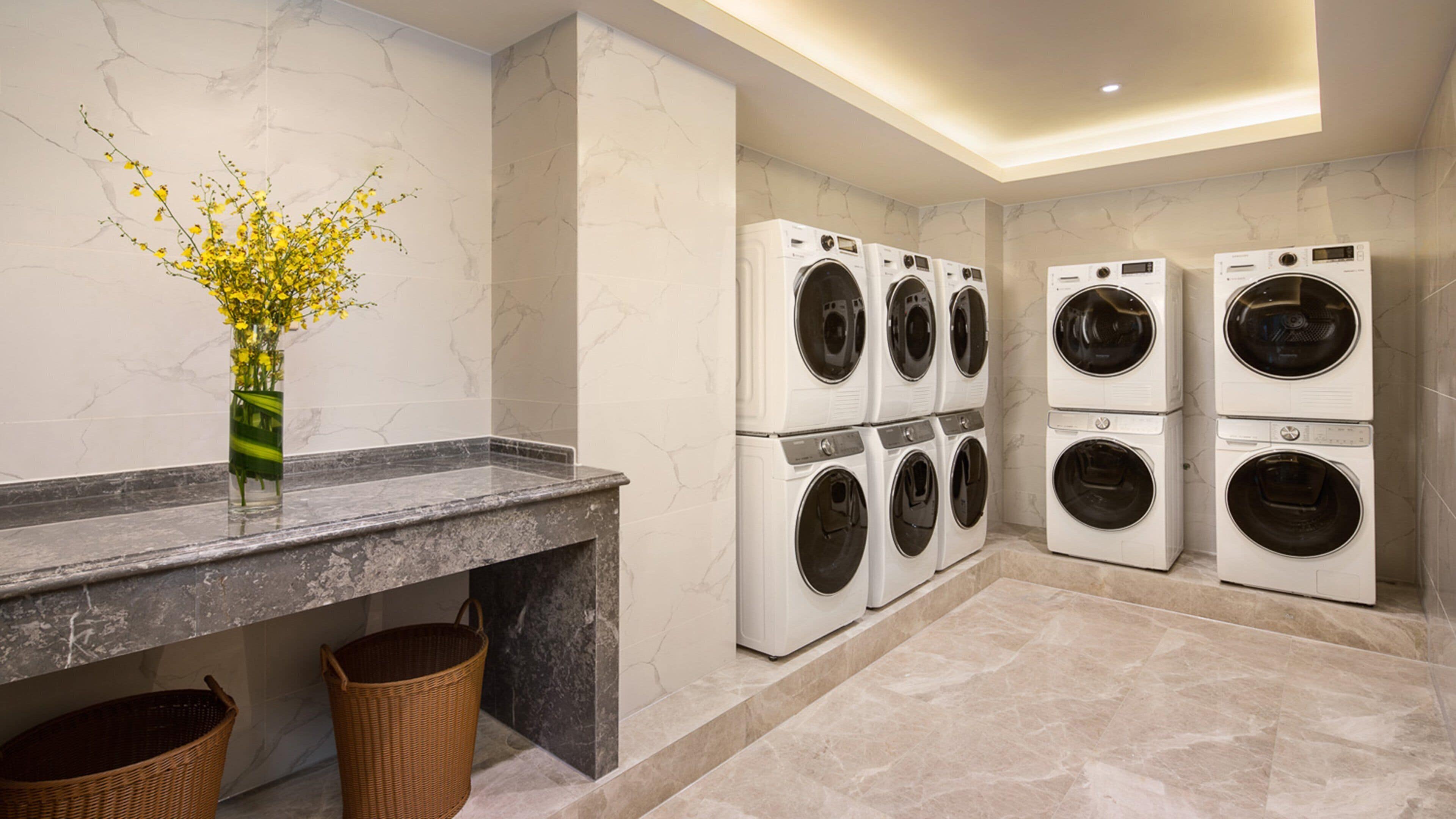 A modern laundry room with four stacked front-loading washing machines, a marble-tiled wall, a marble countertop, and a vase with yellow flowers.