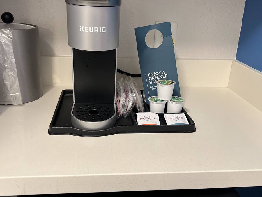 Una cafetera Keurig con una tarjeta azul, dos cápsulas de café y una taza desechable sobre una encimera blanca.