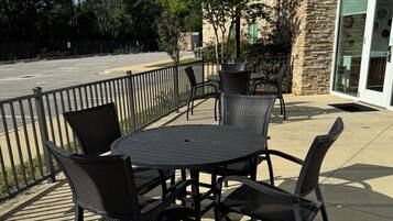 An outdoor patio with a round table and four chairs, a stone building in the background, and a parking lot.
