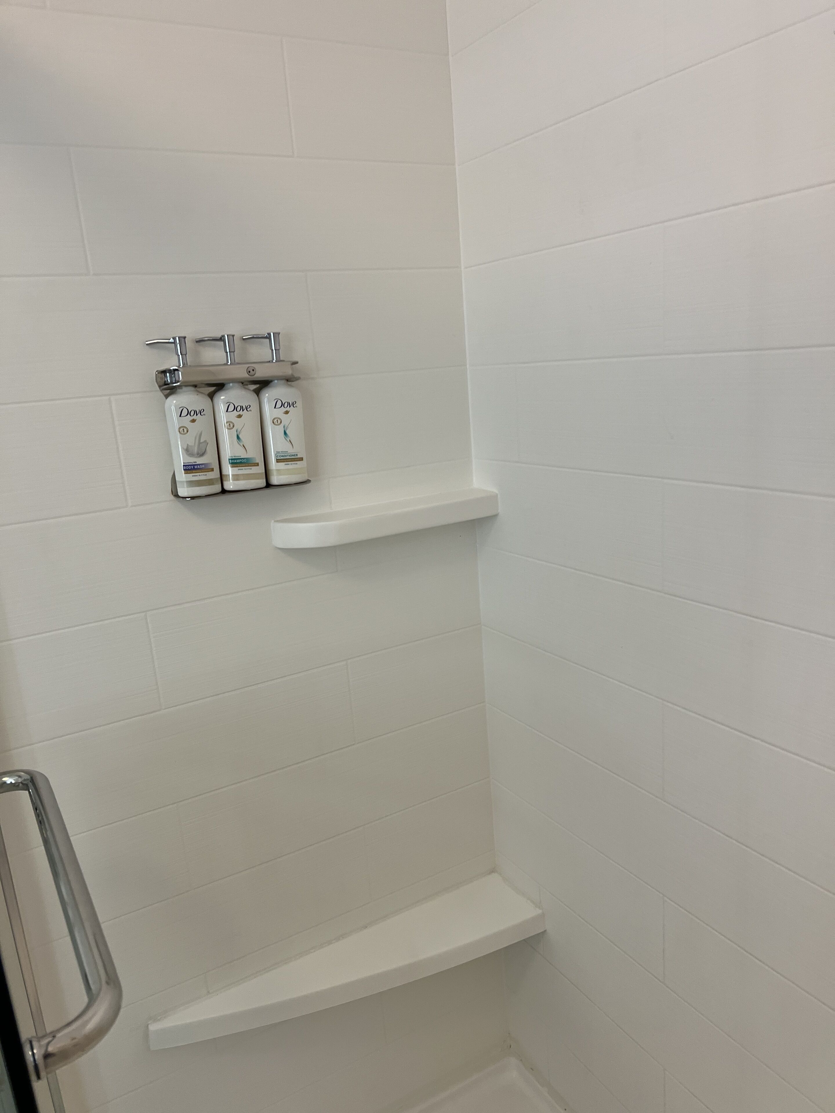 A shower area with white tiles, a metal showerhead, and three Dove body wash bottles on a shelf.