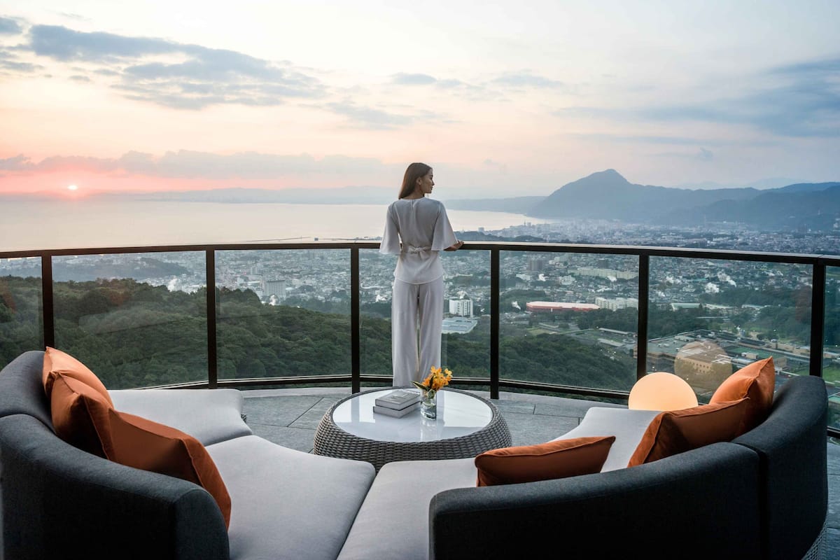 A person standing on a balcony overlooking a cityscape with mountains in the distance.