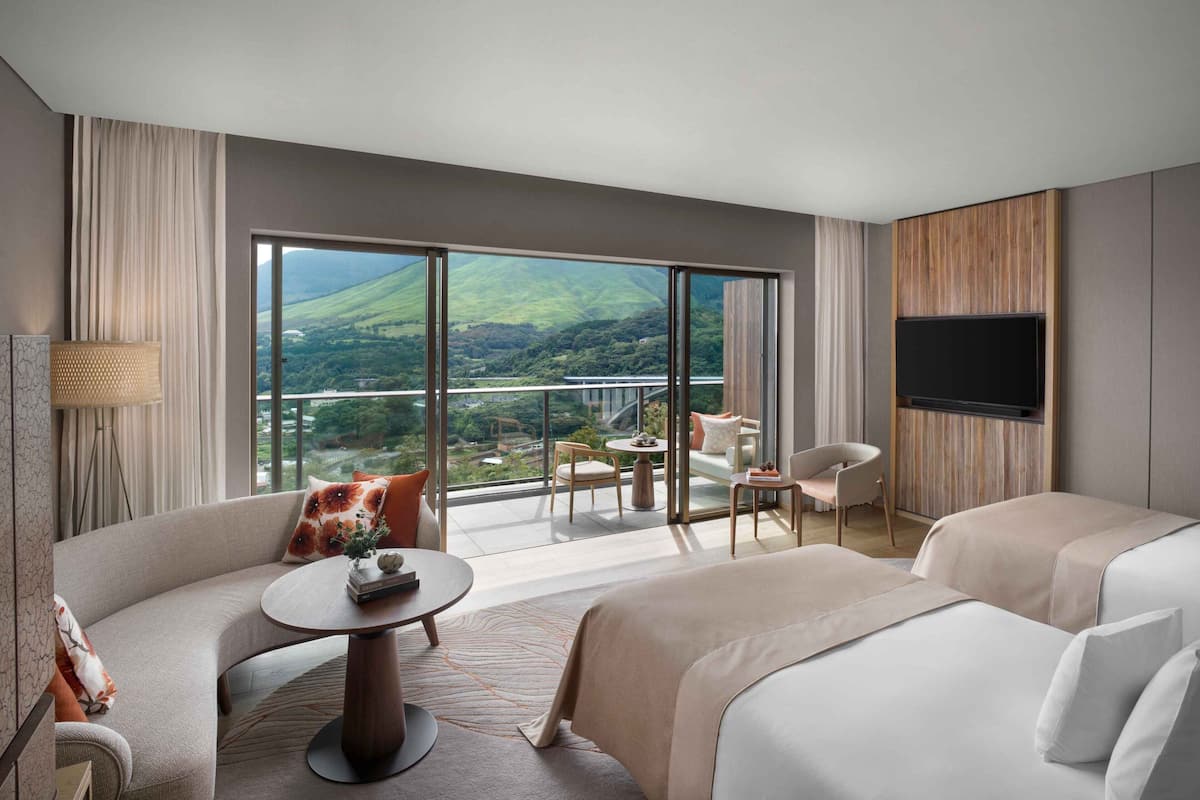 A hotel room with a large window, a sofa, a bed, a TV, and a balcony with a view of the countryside.