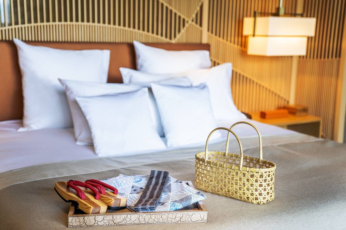 A neatly made bed with white bedding and a headboard, a small wooden tray with slippers and a striped item, and a woven basket on the bed.