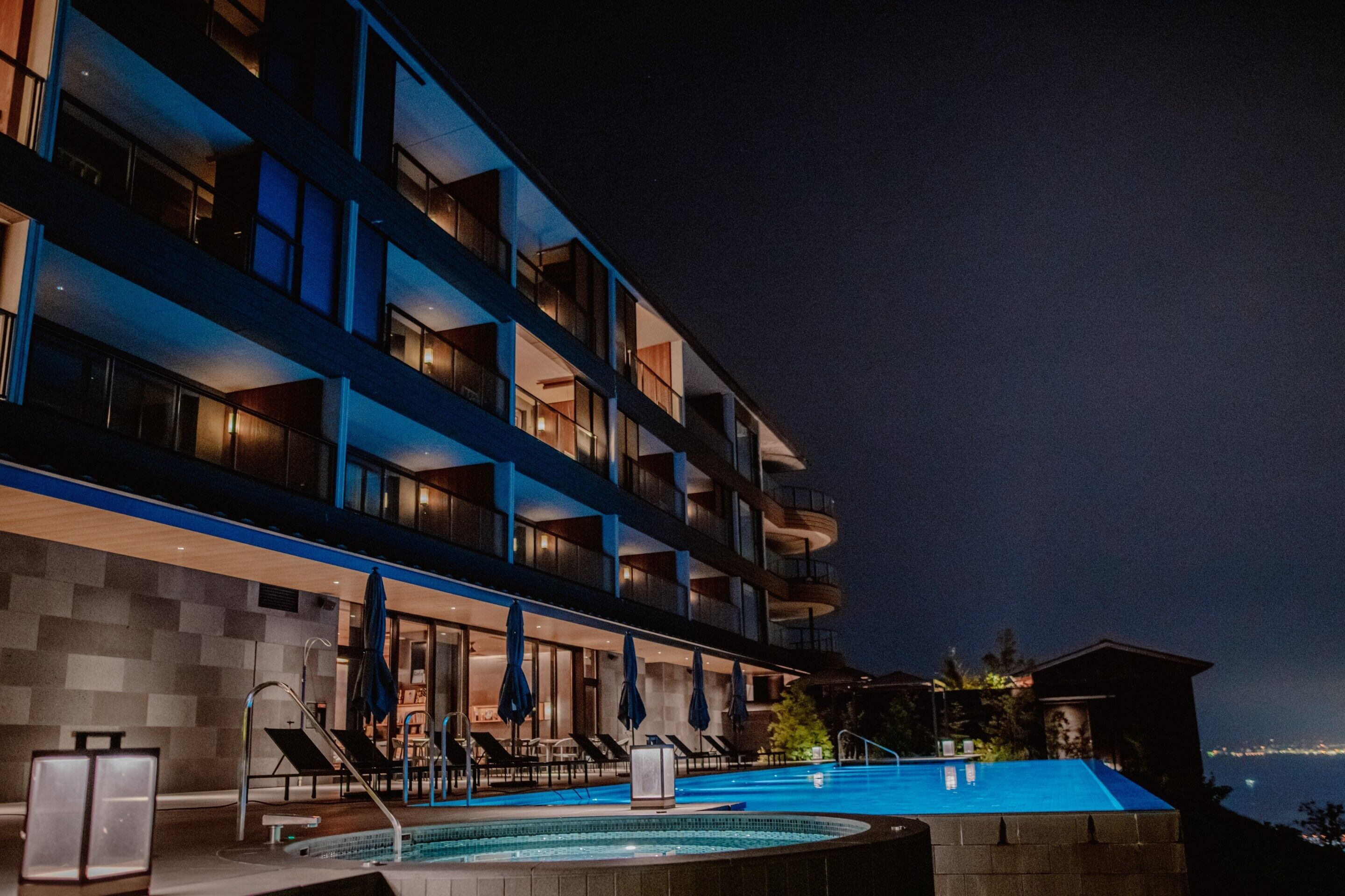 A modern hotel with a pool, multiple balconies, and a view of the sea at night.