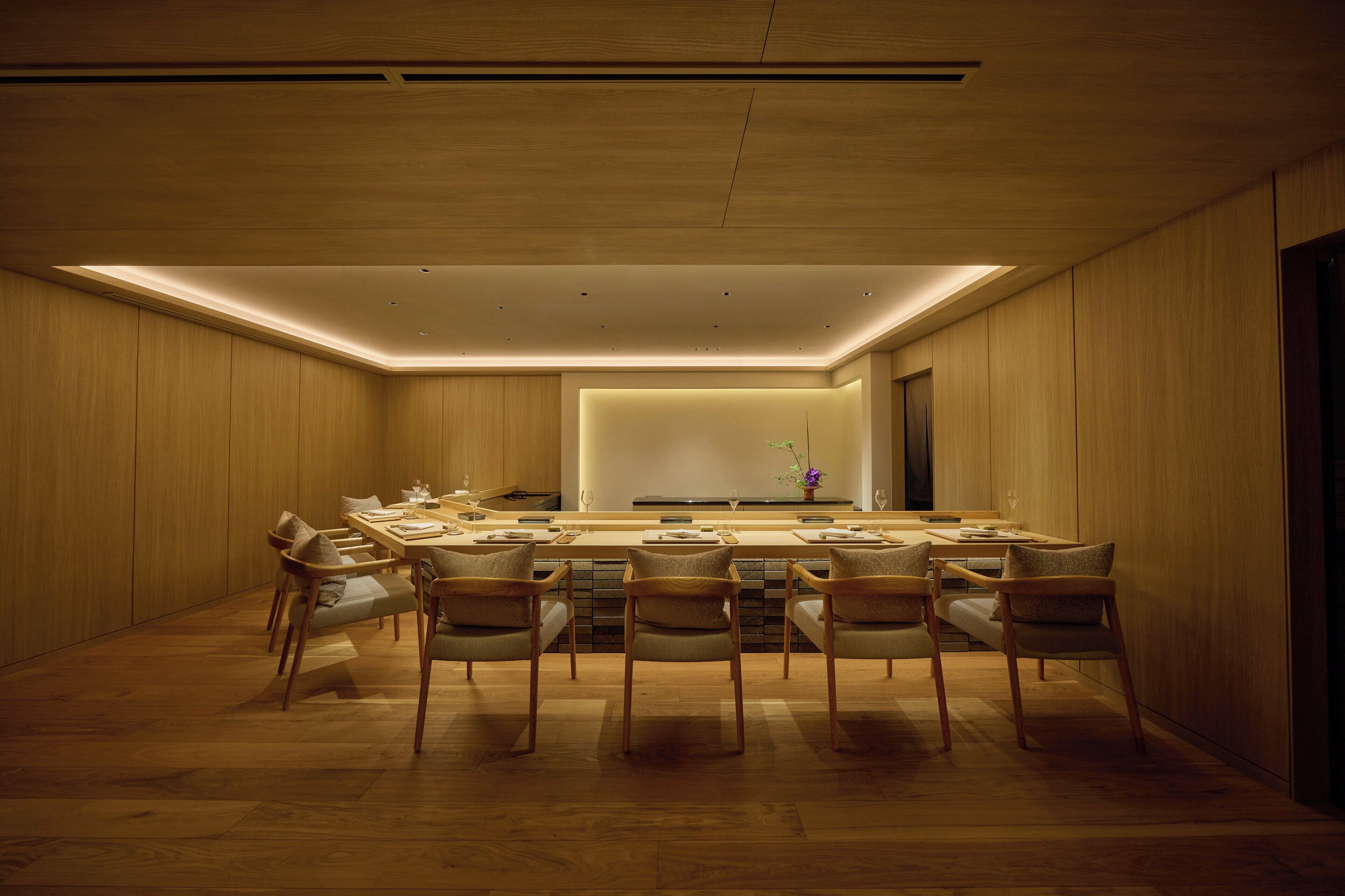 A modern dining area with a long table and six chairs, wooden paneling on the walls, and recessed lighting.