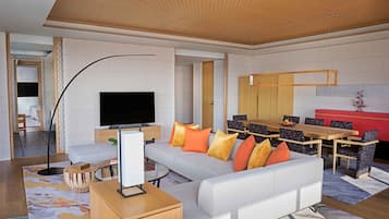A modern living room with a large flat-screen TV, a grey sofa with orange and yellow cushions, a wooden dining table with chairs, and a kitchen area in the background.