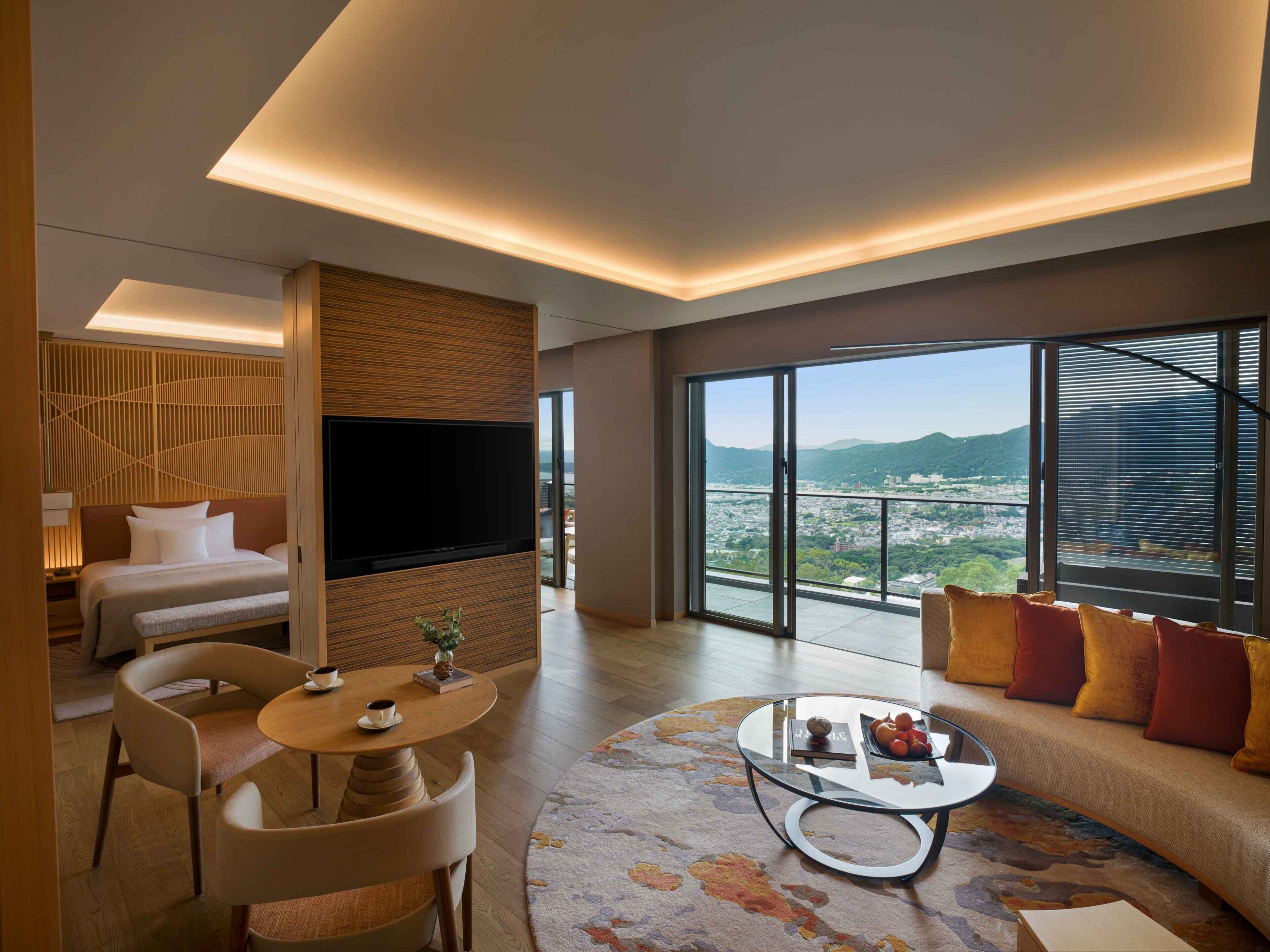 Una moderna camera d'hotel con una grande TV, un divano, un tavolino rotondo e un balcone con vista sulla città.