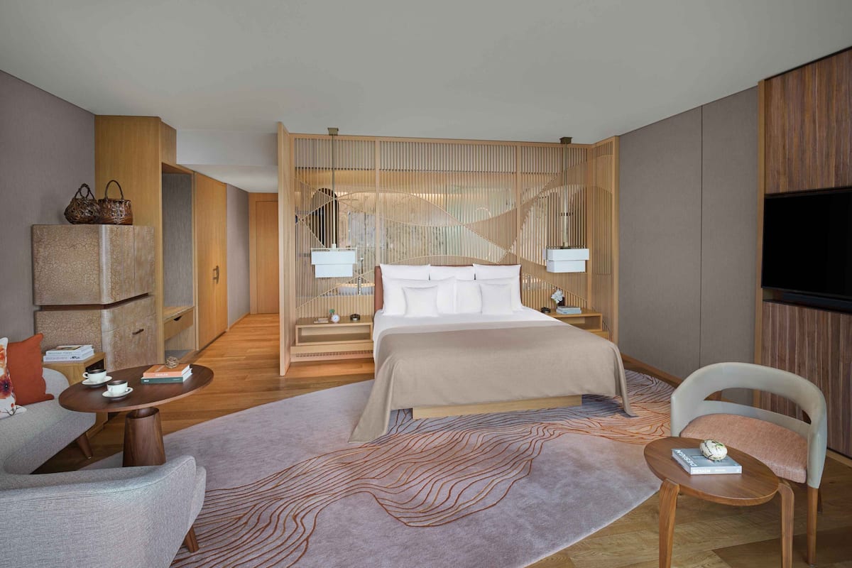 A modern hotel room with a large bed, a TV, a wooden partition, and a seating area with a sofa and a chair.
