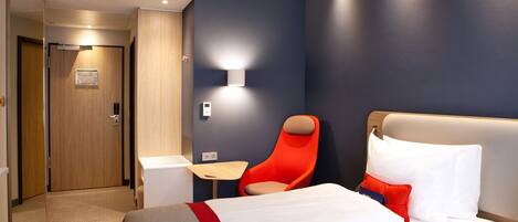 A modern hotel room with a bed, a red chair, a small table, and a wall-mounted light.