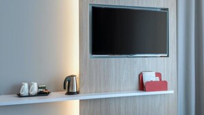 A modern hotel room with a mounted flat-screen TV on a wooden wall, a white shelf with a kettle, cups, and a red folder. - Holiday Inn Express Mulheim - Ruhr by IHG (Muelheim an der Ruhr)