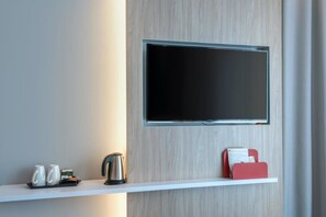 Une chambre d’hôtel moderne avec un téléviseur à écran plat fixé au mur, un mur en bois, une étagère blanche avec une bouilloire, des tasses et un dossier rouge.