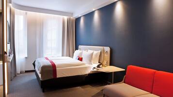 A hotel room with a bed, a bedside table with a phone, a red sofa, and a window with curtains.
