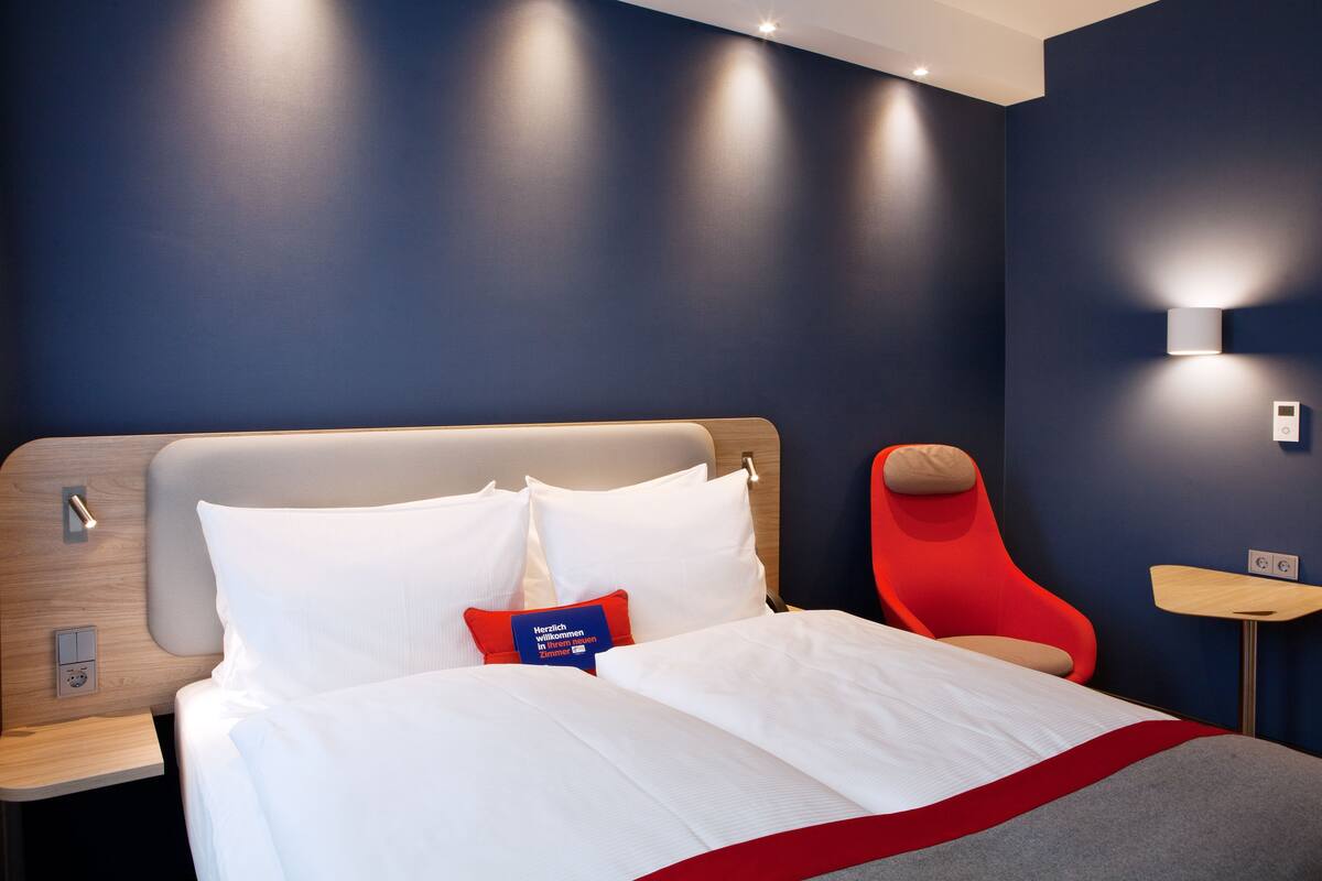 A modern hotel room with a large bed, a red chair, and a small table.