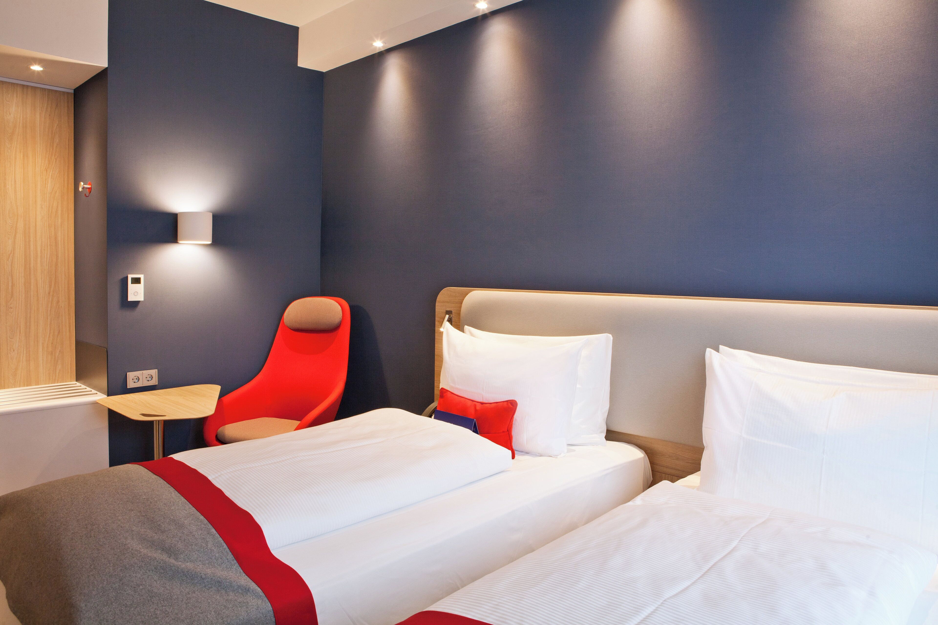 Quarto de hotel com duas camas, uma cadeira vermelha, uma mesa pequena e uma luminária na parede.