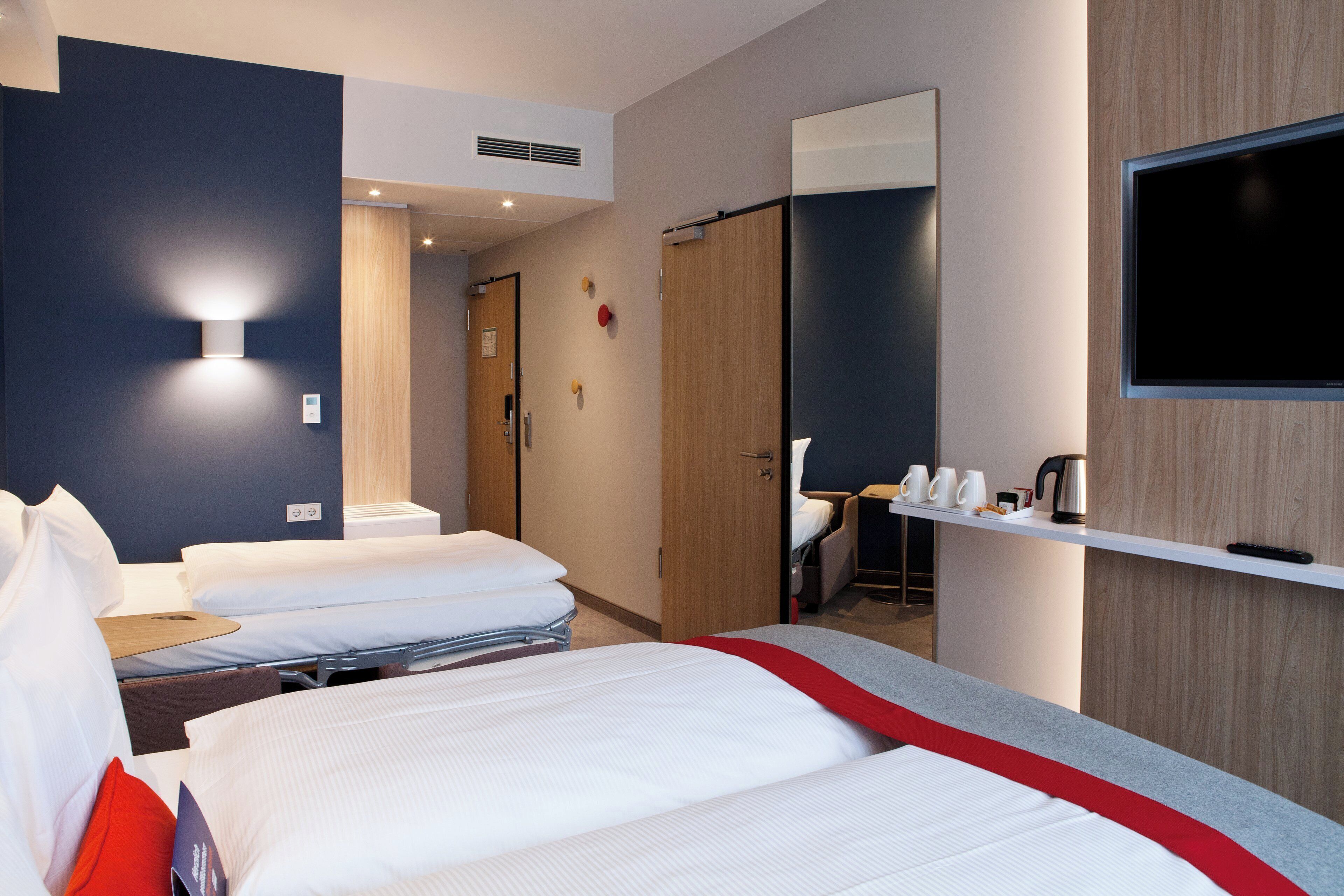 A hotel room with two beds, a TV, a desk with a chair, and a shelf with cups and a kettle.