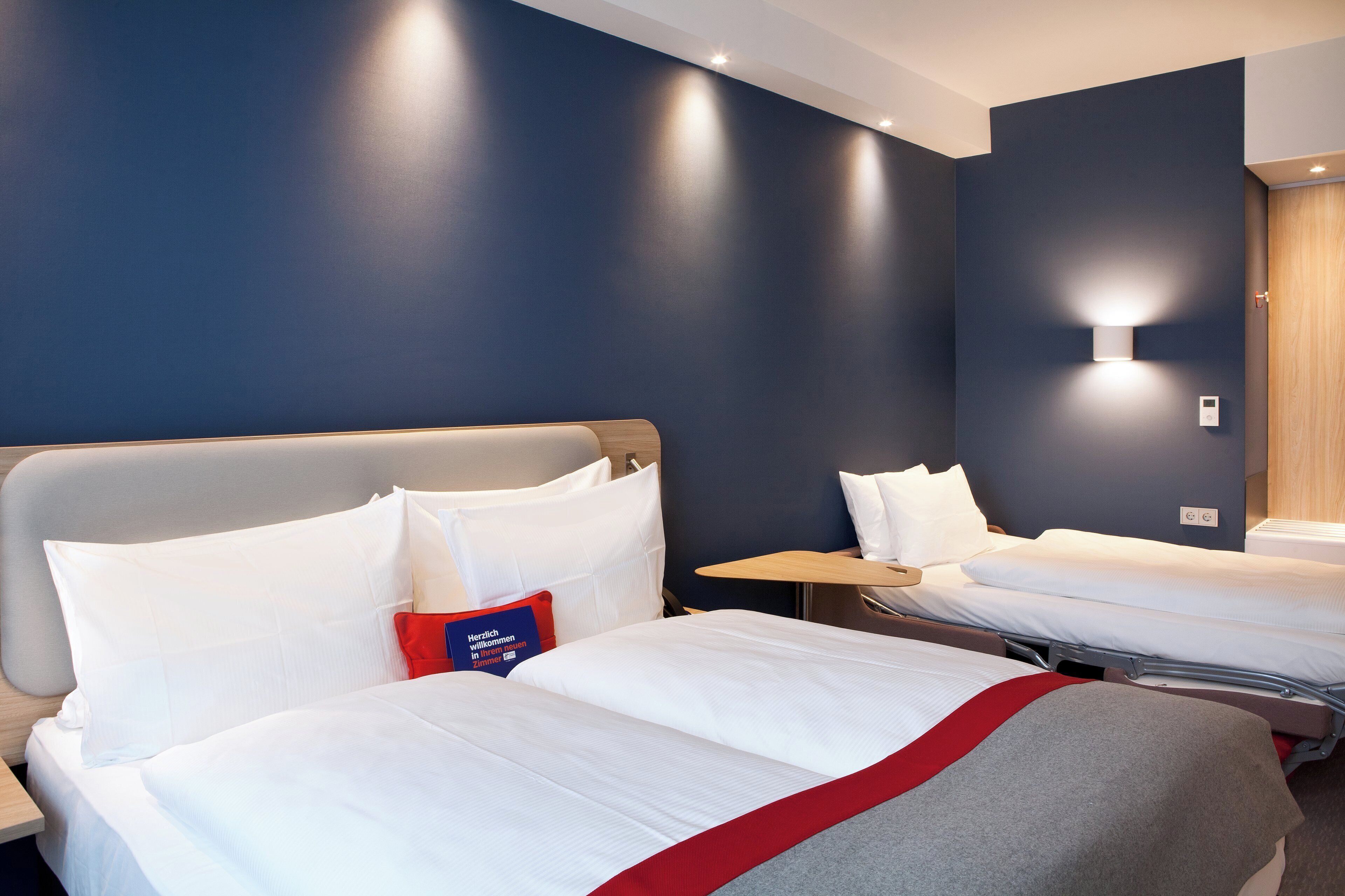 A hotel room with two beds, a bedside table, a wall-mounted light, and a thermostat.