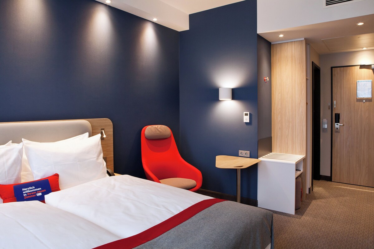 Une chambre d’hôtel avec un lit, une chaise rouge, une petite table et une porte en bois.