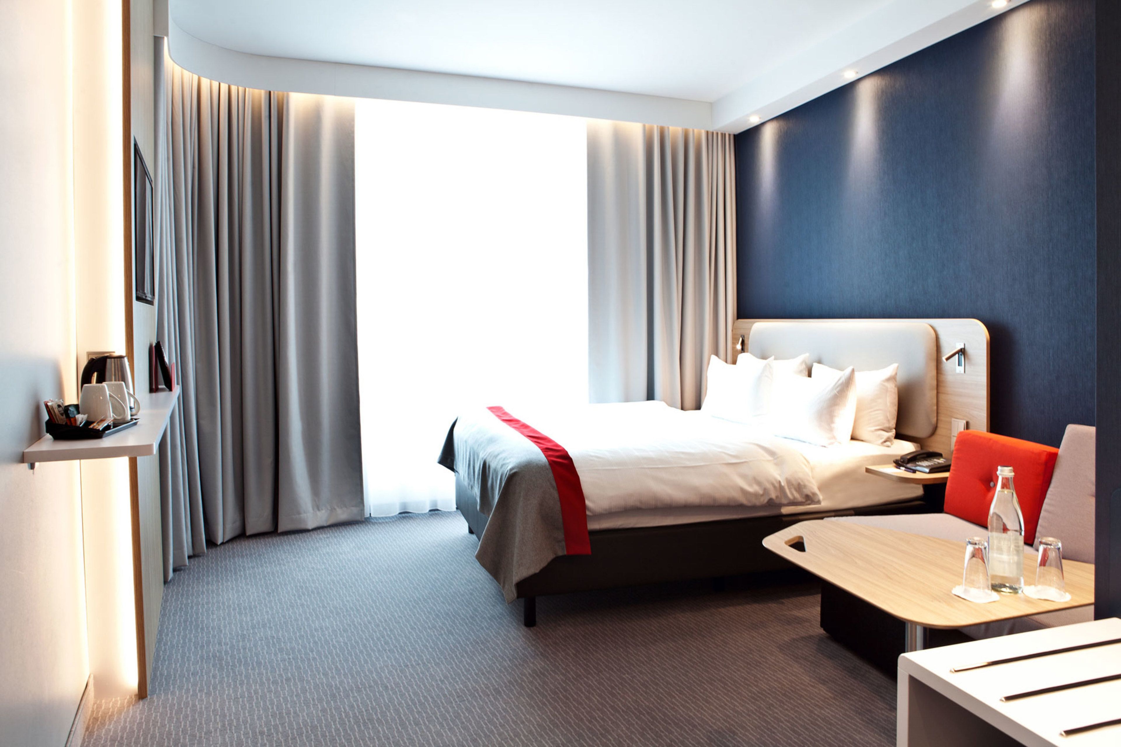 A modern hotel room with a large bed, a bedside table, a small table with glasses, and a view of the outdoors.