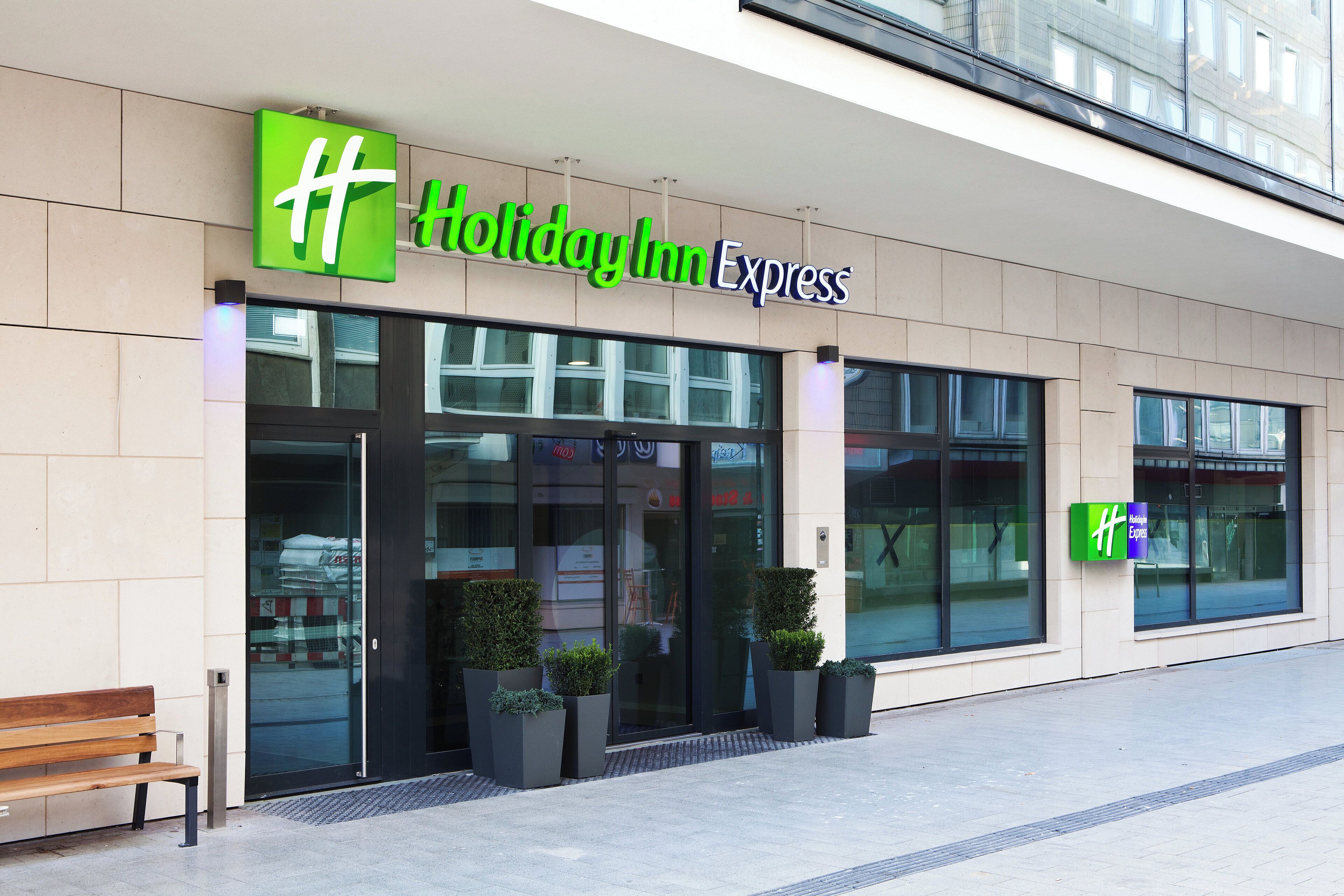 Fachada de un hotel Holiday Inn Express con amplios ventanales, una banca en el exterior y una vista despejada de la entrada.