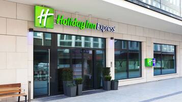 Fachada de un hotel Holiday Inn Express con amplios ventanales, una banca en el exterior y una vista despejada de la entrada.