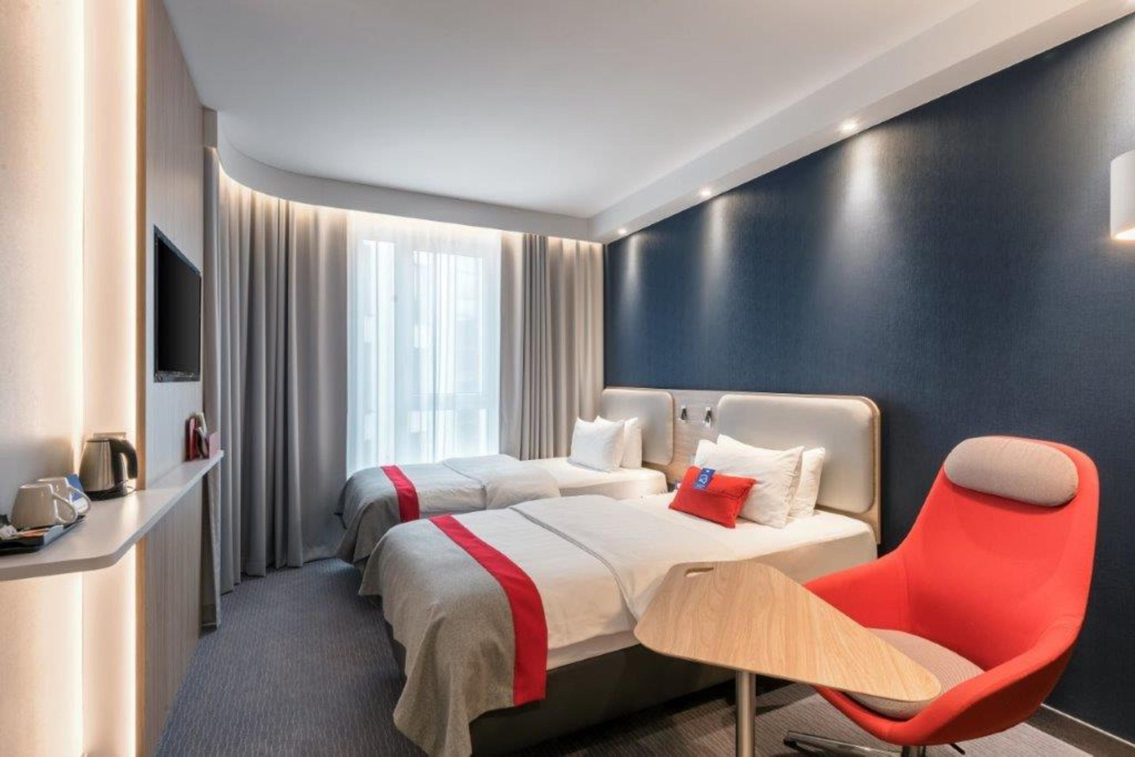 A hotel room with two beds, a red chair, a small table, a TV, and a window with curtains.