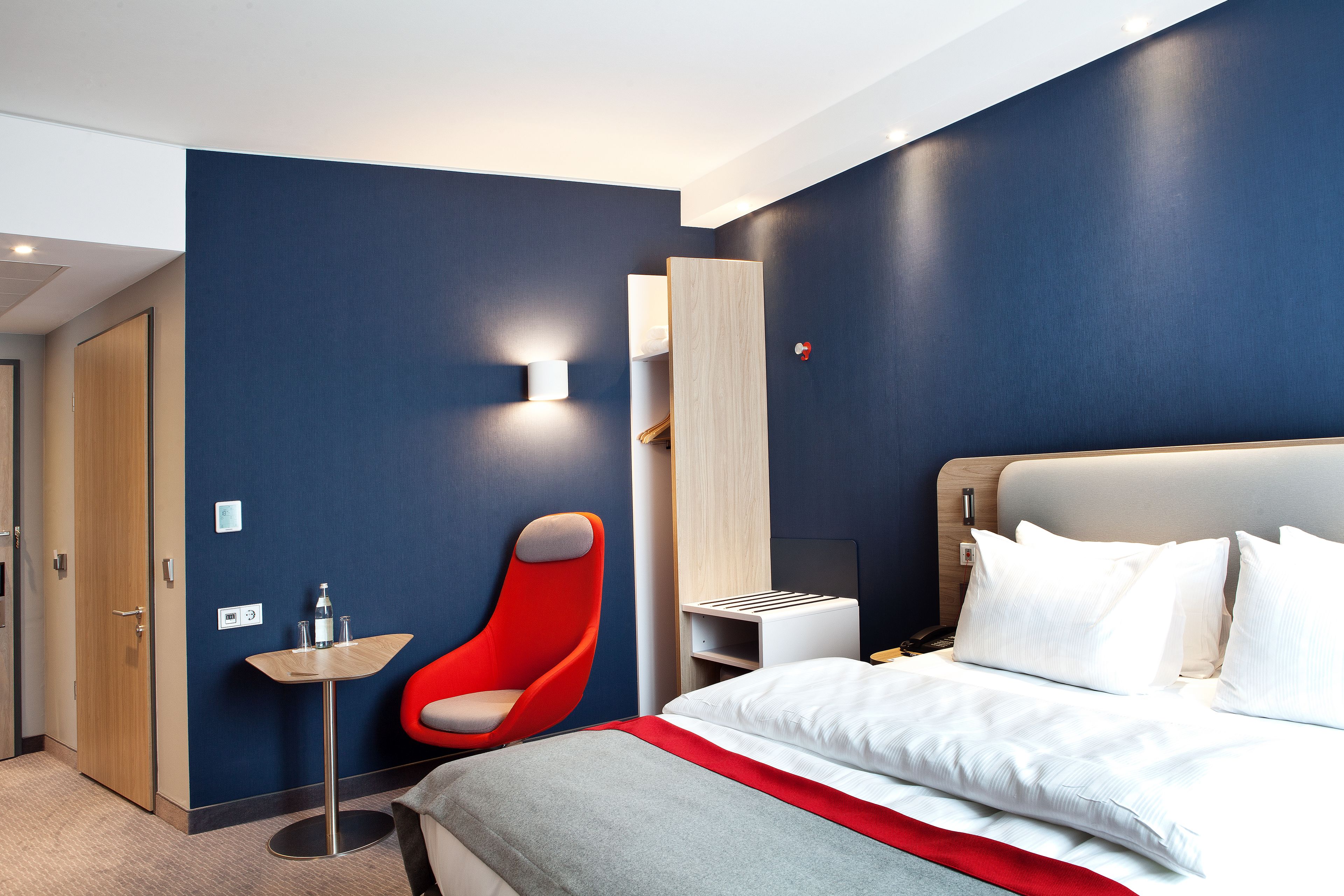 Una habitación de hotel moderna con una cama, una silla roja, una mesita pequeña y una mesita de noche.