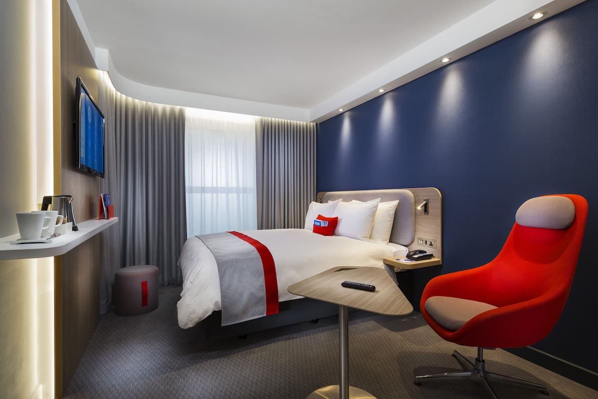 A hotel room with a bed, a red chair, a TV, and a small table.