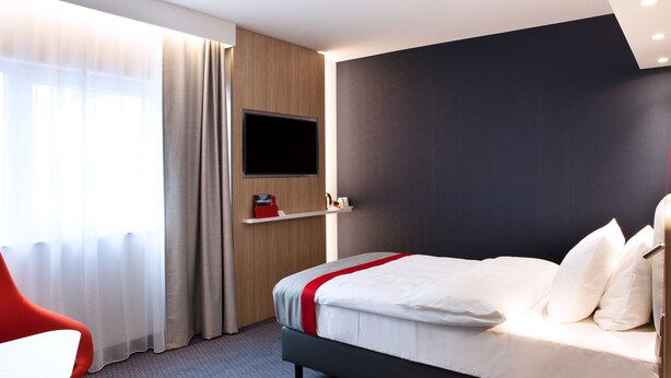 A modern hotel room with a bed, a TV mounted on the wall, a red chair, and a window with curtains. - Holiday Inn Express Oberhausen by IHG (Oberhausen)